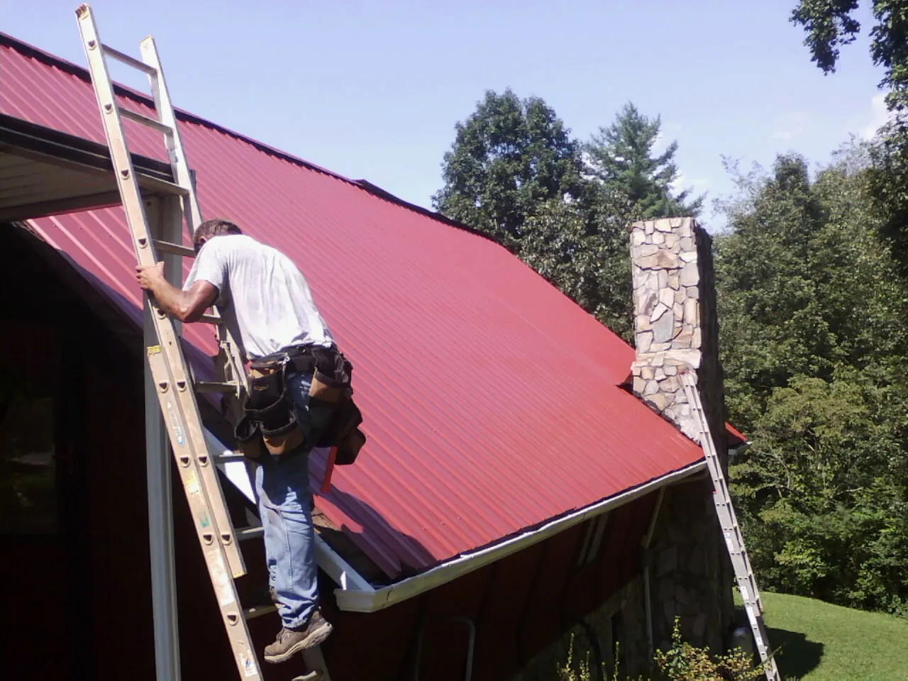 Roofer on a ladder beside a red metal roof and stone chimney — Soffit & Fascia Repair by Roofing Brothers in Du Quoin